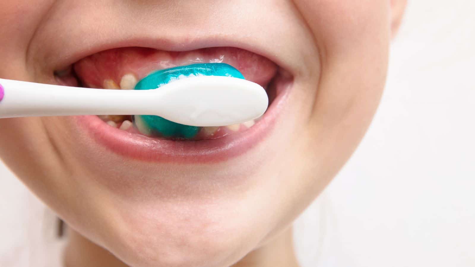 A close-up of a child's mouth smiling while brushing their teeth with a toothbrush covered in blue toothpaste. Some baby teeth and gaps are visible.