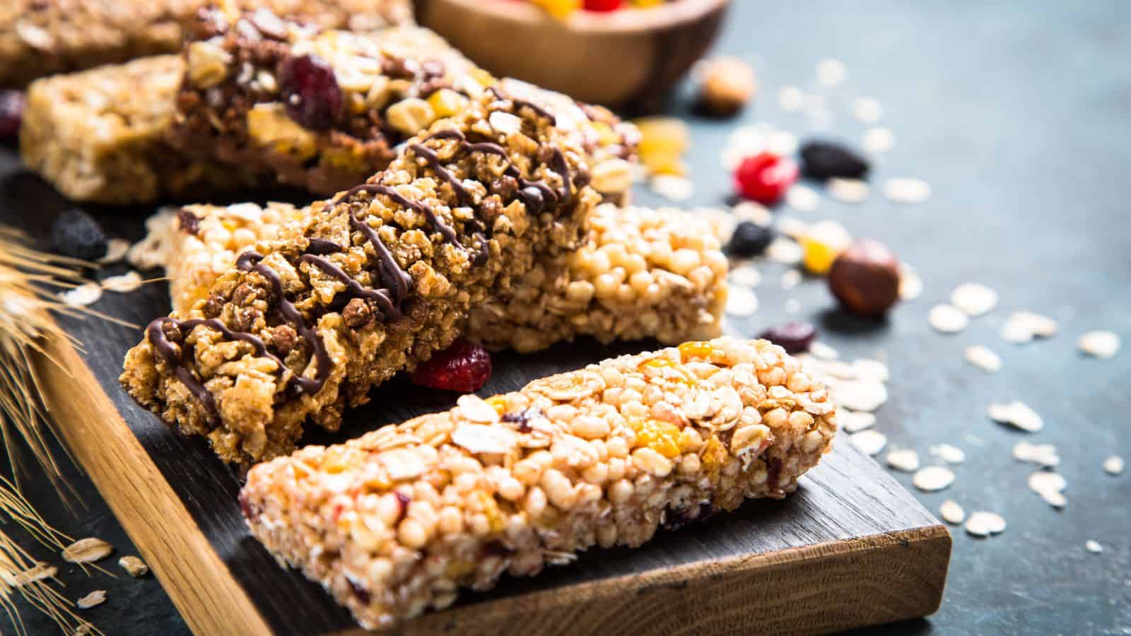 Several granola bars with various toppings, such as chocolate drizzle, nuts, and dried fruit, are arranged on a wooden board. Loose oats and bits of dried fruit are scattered around the board.