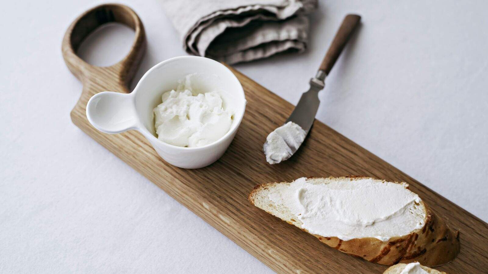 A slice of bread with a layer of spread next to a knife and a small white bowl of the same spread, all placed on a wooden cutting board with a folded cloth in the background.