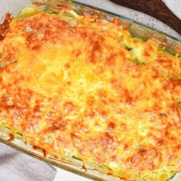 A baked casserole in a rectangular glass dish features a golden-brown, melted cheese topping with visible edges of sliced vegetables underneath. A wooden utensil and a folded gray cloth are nearby on a white surface.