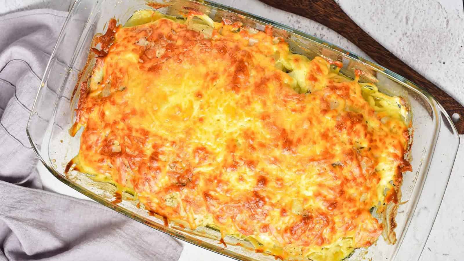 A rectangular glass baking dish filled with a baked casserole topped with melted, browned cheese sits on a white surface beside a folded gray cloth and a wooden utensil.