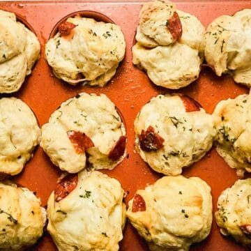 A muffin tin filled with twelve baked savory muffins, some with visible pieces of pepperoni and herbs on top, sits on a light surface. The muffins are golden brown and unevenly shaped.