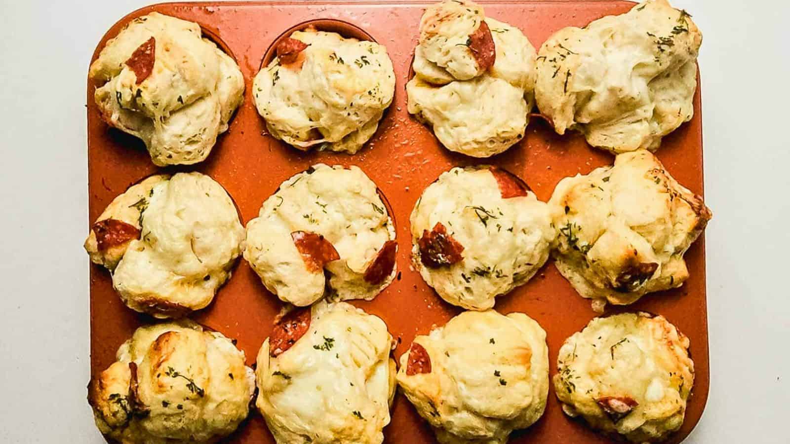 Three savory muffin-shaped pastries with melted cheese and slices of pepperoni on top are stacked on a plate. A cup of sauce and more pastries are visible in the blurred background.