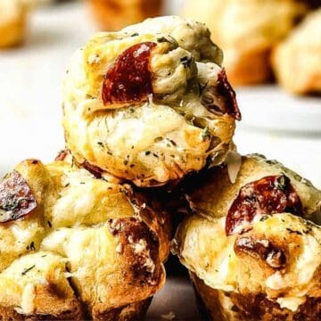 Three savory muffins topped with melted cheese and pepperoni slices are stacked on a white plate. A small bowl of sauce and more muffins are visible in the blurred background.