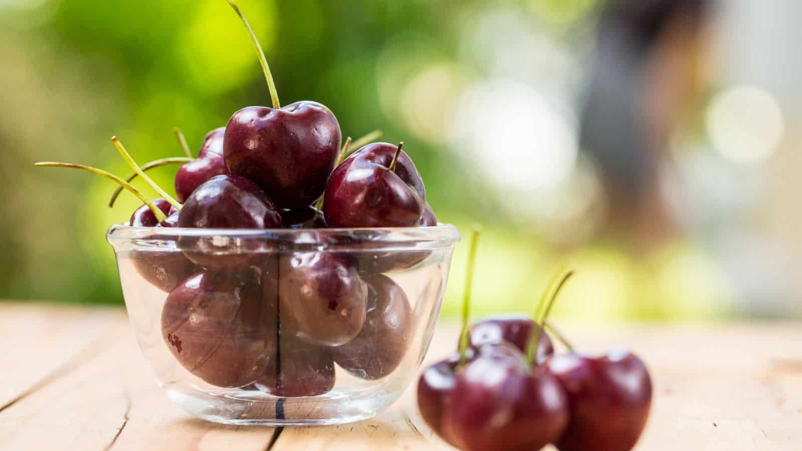 A clear glass bowl filled with dark red cherries sits on a wooden surface. Several cherries with stems are lying next to the bowl. The background is blurred with green and light colors.