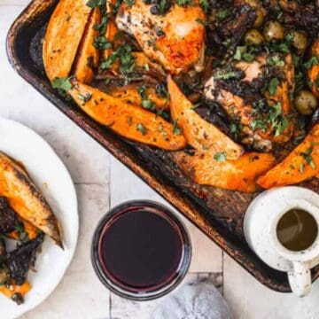 A baking tray with roasted chicken, sweet potato wedges, olives, and herbs is on a table next to a plate of the same food, a glass of red wine, and a small jug of sauce.