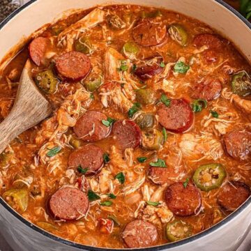 A large pot of gumbo with sliced sausage, shredded chicken, okra, and herbs in a thick, reddish-brown broth. A wooden spoon rests inside the pot and a plate of cornbread is visible nearby.
