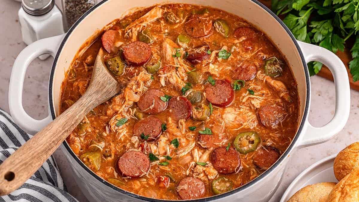 A pot of gumbo with sausage, chicken, and sliced vegetables. A wooden spoon rests inside the pot. Salt and pepper shakers, parsley, and bread rolls are visible in the background.