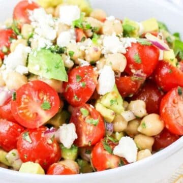A white bowl filled with a salad made of halved cherry tomatoes, chickpeas, diced avocado, chopped red onion, cilantro, and crumbled white cheese, placed on a table with a few loose cherry tomatoes nearby.