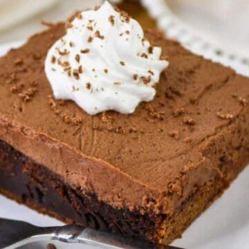 A square slice of chocolate mousse cake topped with a swirl of whipped cream and chocolate shavings sits on a white plate with a fork and a few chocolate chips nearby.