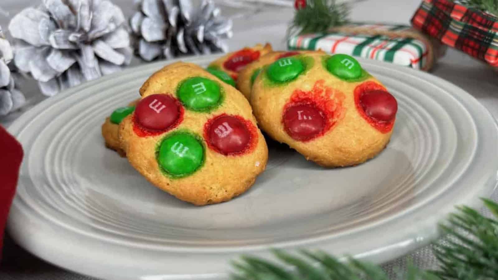 Two cookies topped with red and green chocolate candies on a gray plate, surrounded by pinecones and wrapped gifts.