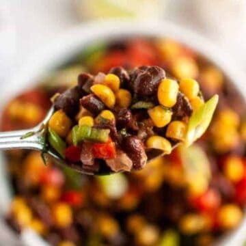 A close-up of a spoon holding a mix of black beans, corn, chopped red peppers, and green onions, with a bowl of the same mixture blurred in the background.