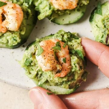 A hand holds a cucumber slice topped with mashed avocado and a cooked shrimp. Other similar cucumber appetizers are arranged on a plate in the background.