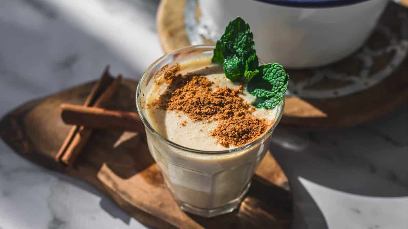A glass filled with a creamy beverage topped with ground cinnamon and fresh mint leaves, placed on a wooden board next to two cinnamon sticks. Sunlight casts shadows on the scene.