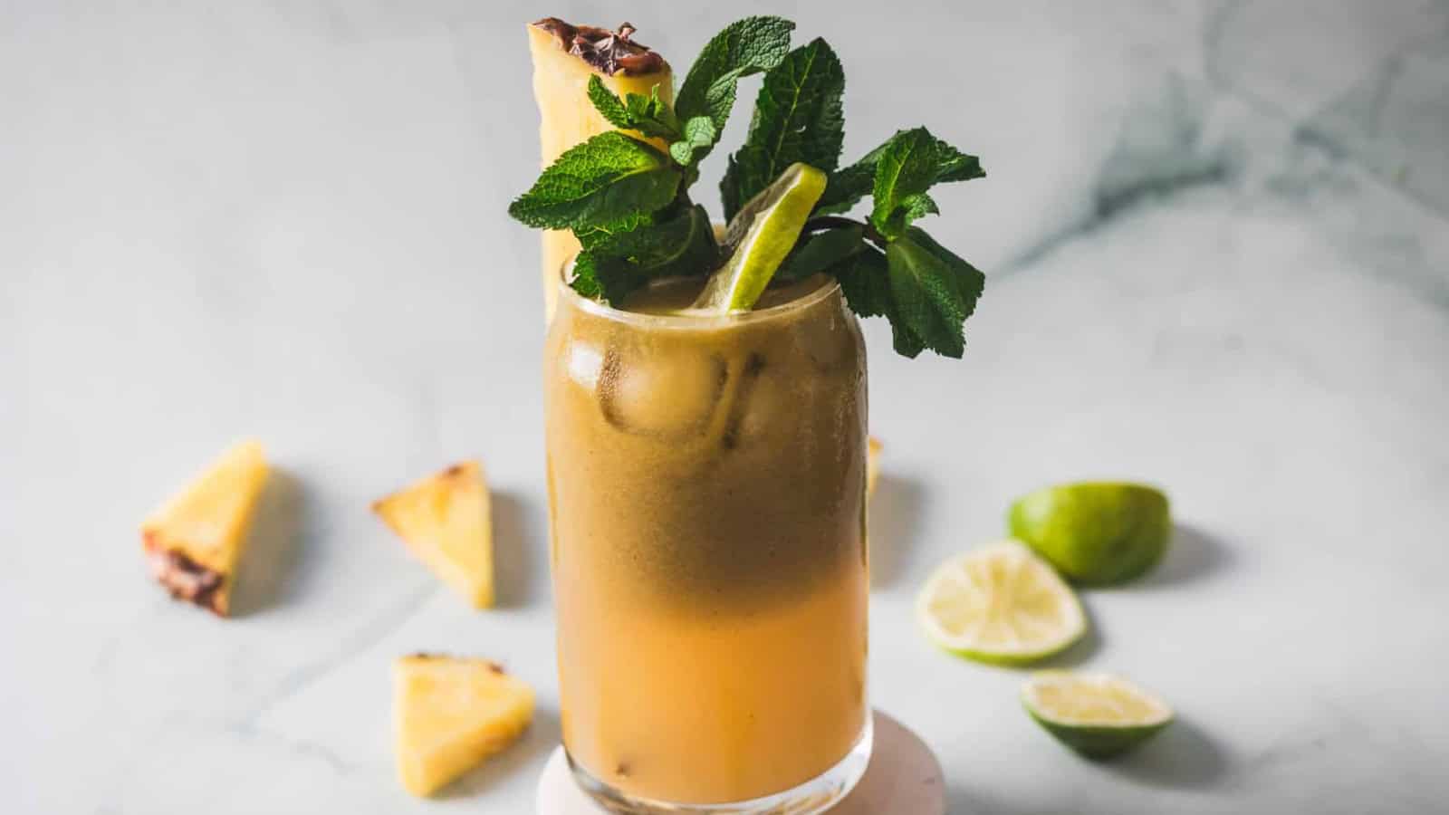 A tall glass of iced brownish-orange beverage garnished with a sprig of fresh mint, a pineapple wedge, and a lime wedge. Slices of pineapple and lime are scattered on a marble surface in the background.