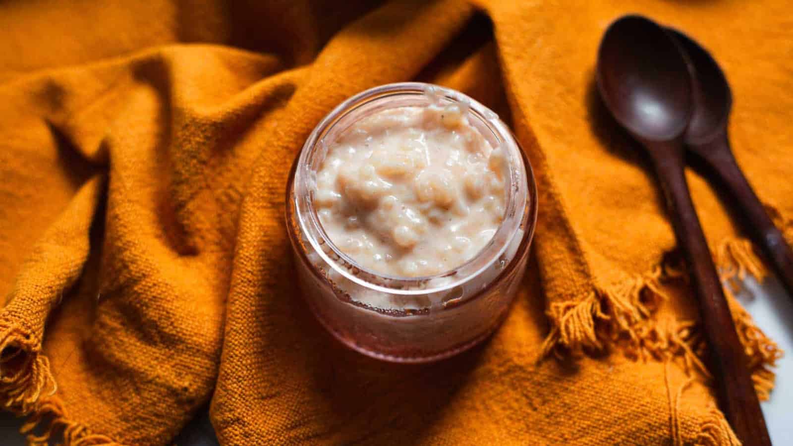 A glass jar filled with rice pudding sits on an orange textured fabric next to two dark wooden spoons.