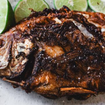A whole fried fish with crispy, browned skin is served on a plate with several lime wedges arranged around it.