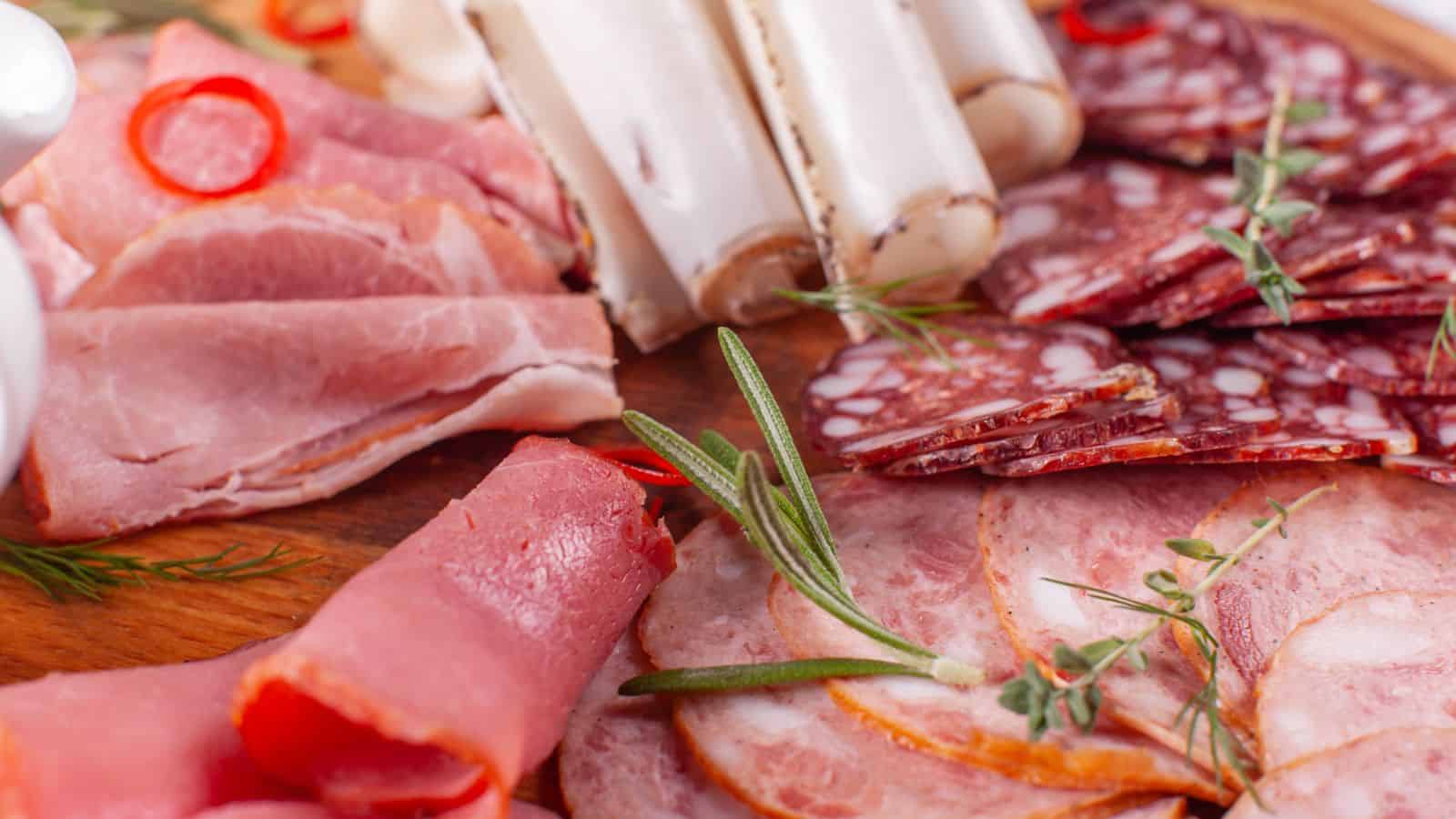 A wooden board with a variety of sliced cured meats, including ham, salami, and sausage, garnished with rosemary, dill, and slices of chili pepper.