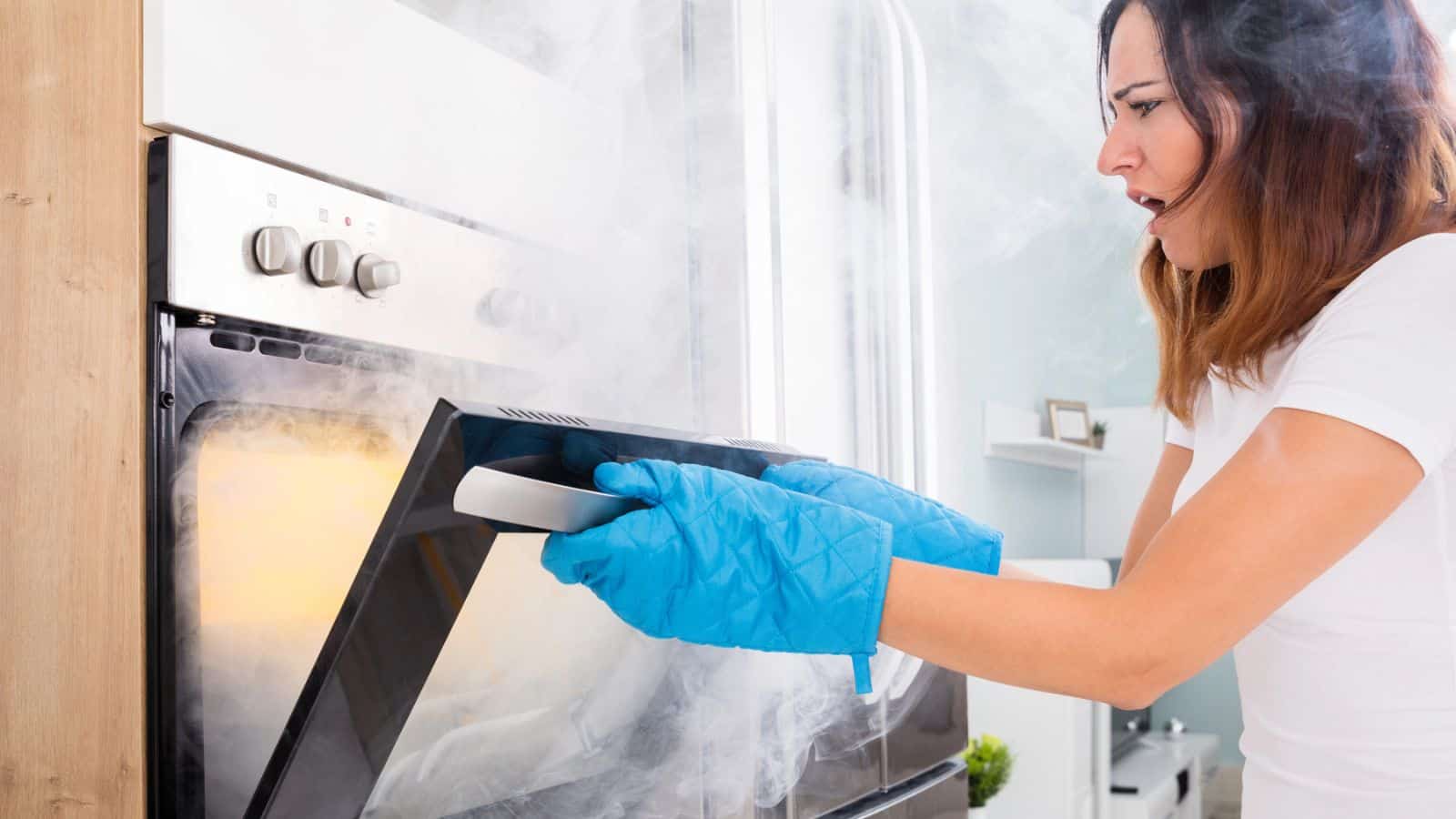A woman wearing blue oven mitts opens an oven filled with smoke. She looks surprised or concerned, standing in a modern kitchen. The oven door is partially open, and the room is visibly smoky.