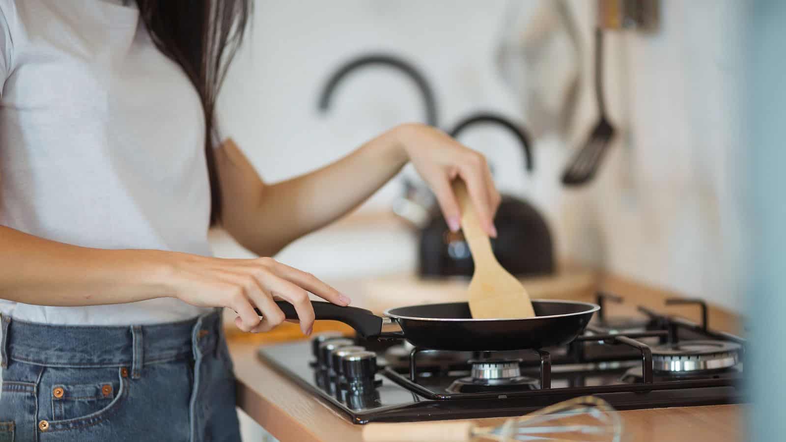 A person wearing a white t-shirt and jeans holds a frying pan and stirs food with a wooden spatula on a gas stove in a kitchen. Kitchen utensils and a kettle are visible in the background.