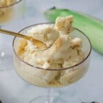 A gold spoon scoops a serving of pale yellow corn ice cream from a glass dish, with a blurred green corn cob in the background on a light surface.