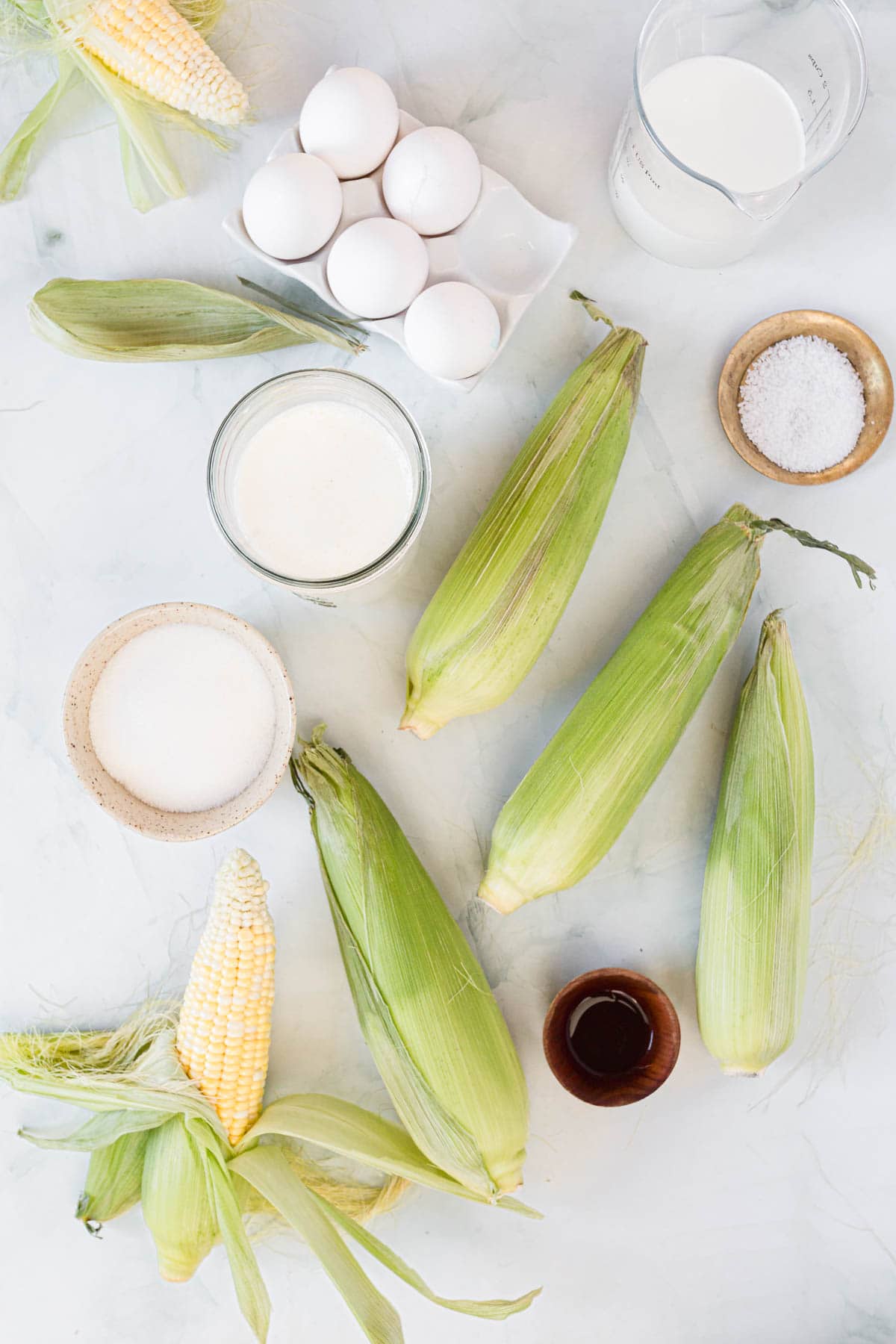 Overhead view of fresh corn on the cob, eggs in a carton, a cup of milk, a cup of cream, a bowl of sugar, a bowl of salt, and a small cup of vanilla extract on a light surface.