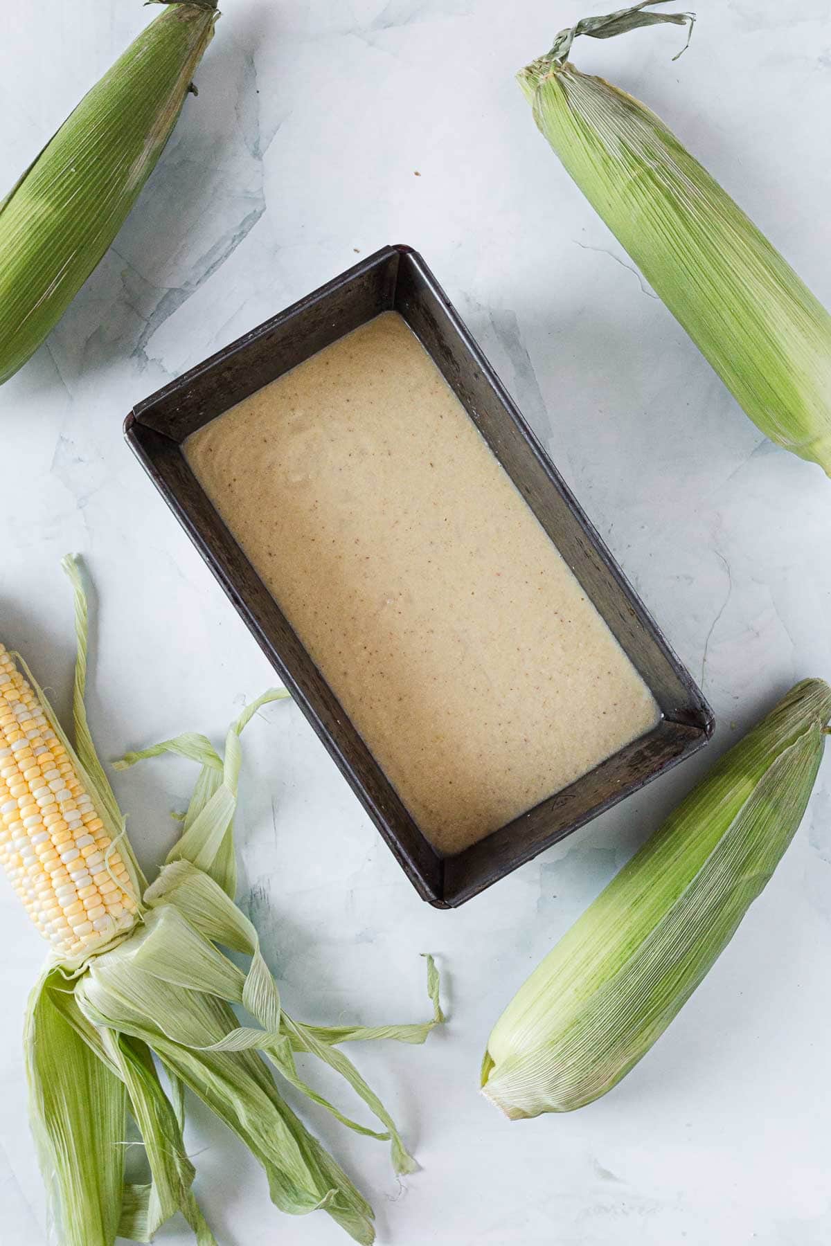 A rectangular metal loaf pan filled with cornbread batter sits on a light surface, surrounded by four ears of corn—perfect inspiration for cornbread or even a unique Corn Ice Cream dessert.