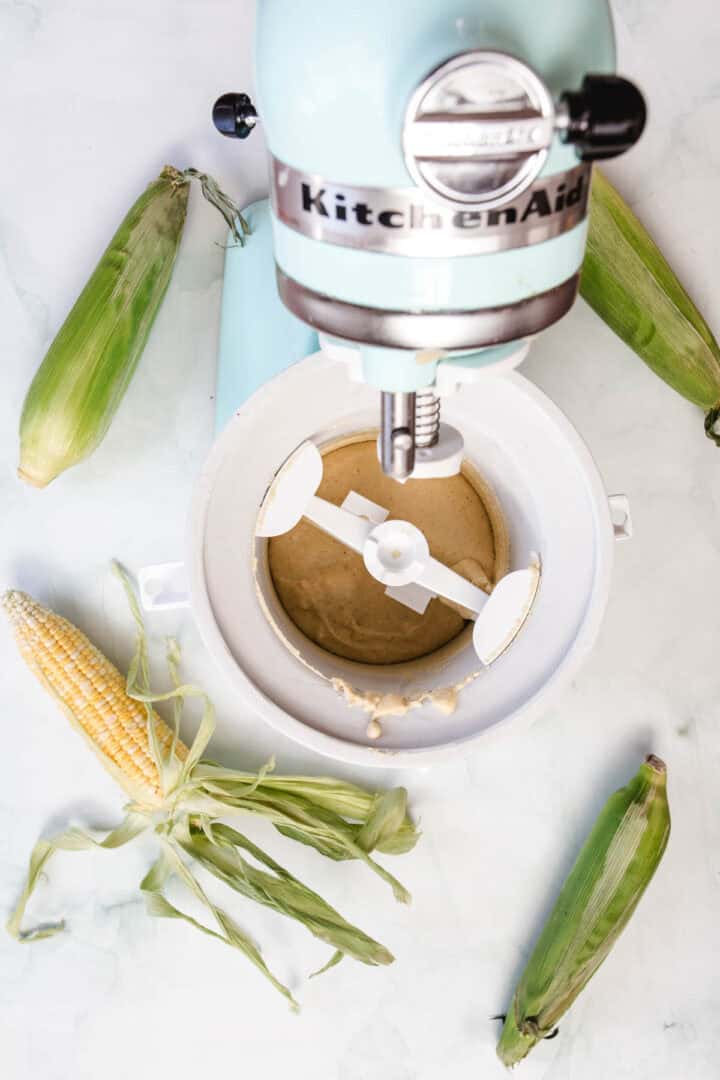 A stand mixer with a corn ice cream attachment contains a light brown mixture. Four ears of corn, three in husks and one partially husked, are arranged around the mixer on a white surface.