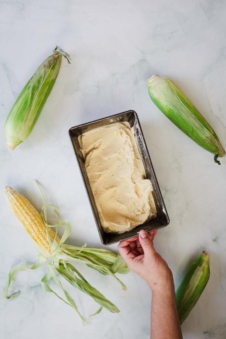 A hand holds a rectangular metal container filled with creamy yellow Corn Ice Cream, surrounded by three ears of corn in husks and one partially husked ear on a white surface.