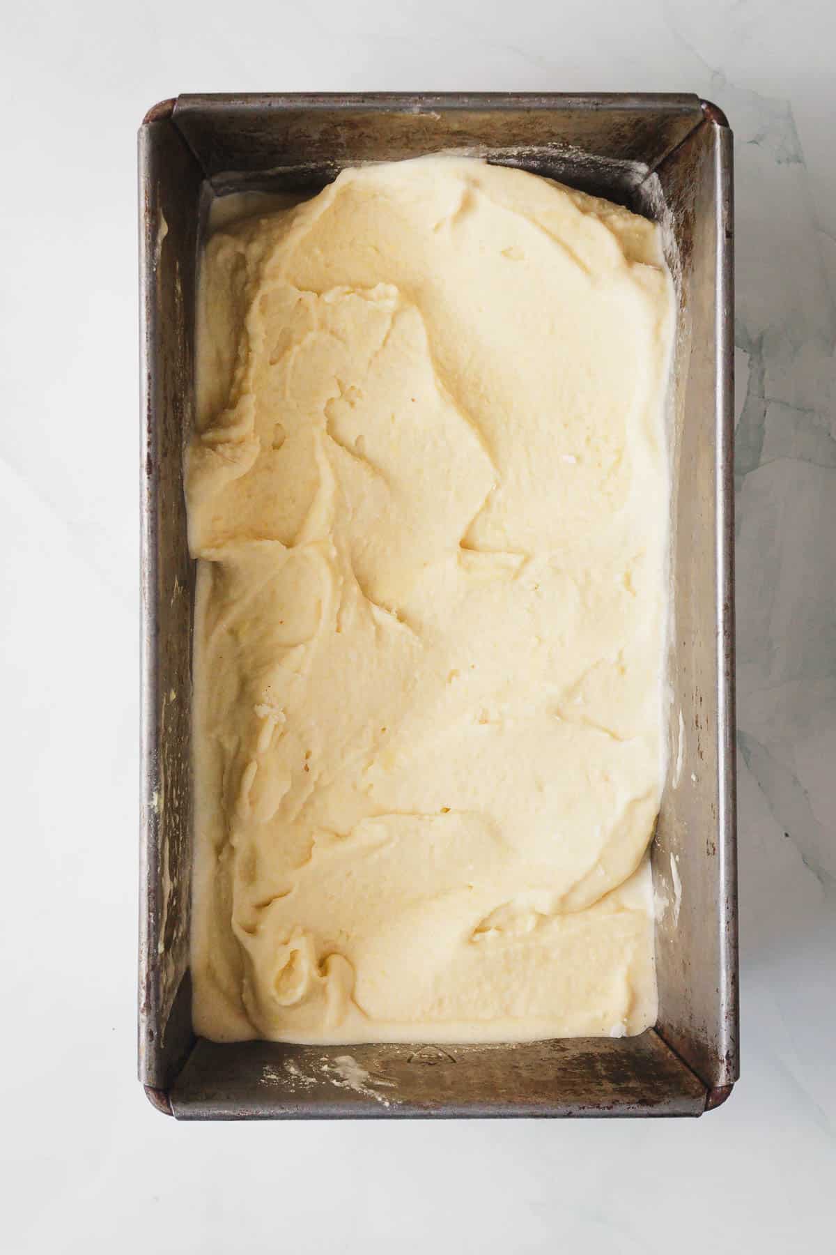 A metal loaf pan filled with unbaked corn ice cream cake batter, spread evenly to the edges, sits on a white marble surface.