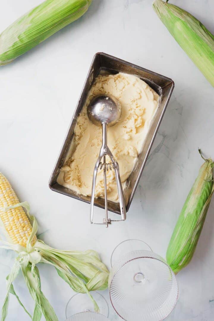 A metal loaf pan filled with pale yellow Corn Ice Cream and a metal scoop, surrounded by whole ears of corn with husks, on a white surface with two empty glass dishes nearby.