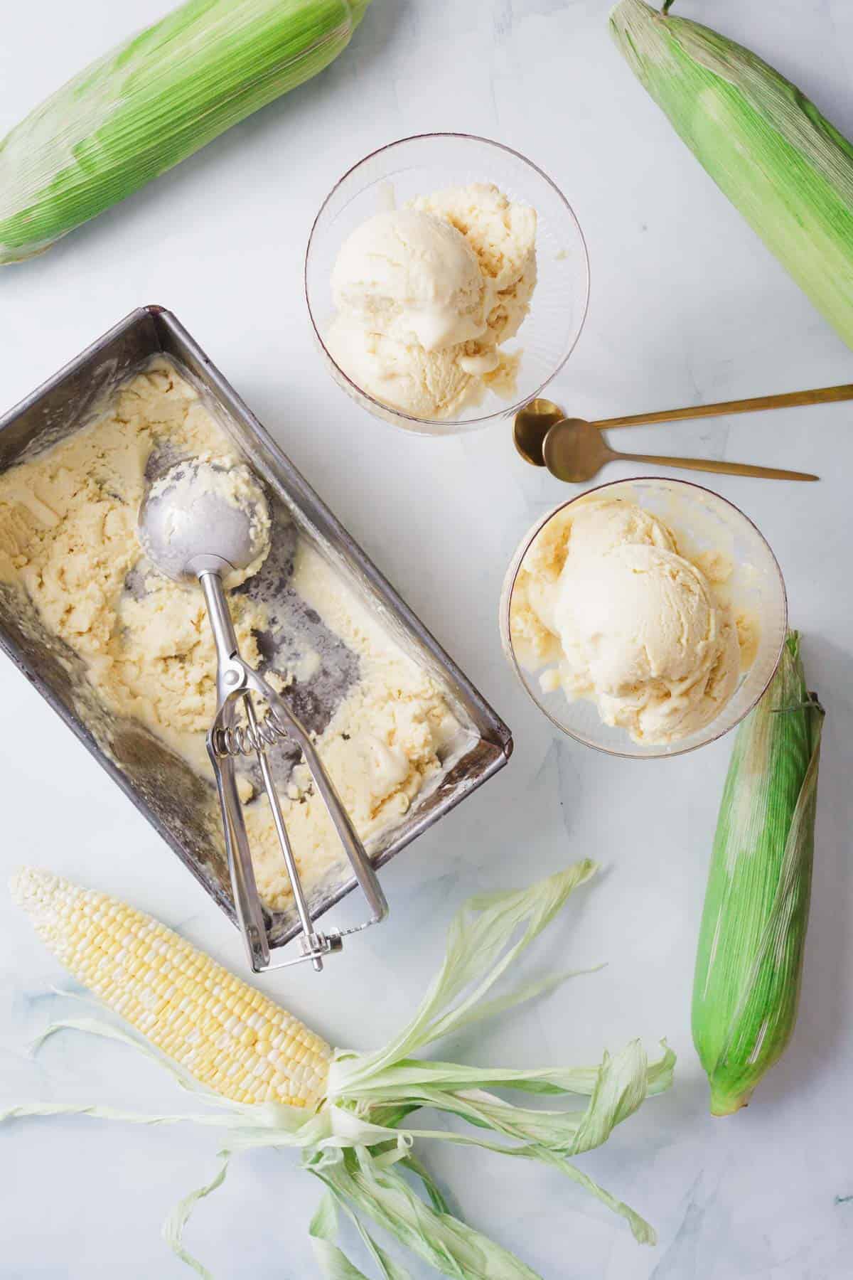 A metal container of corn ice cream with a scoop, two bowls of ice cream, two gold spoons, and four ears of corn—one partially husked—on a white surface.