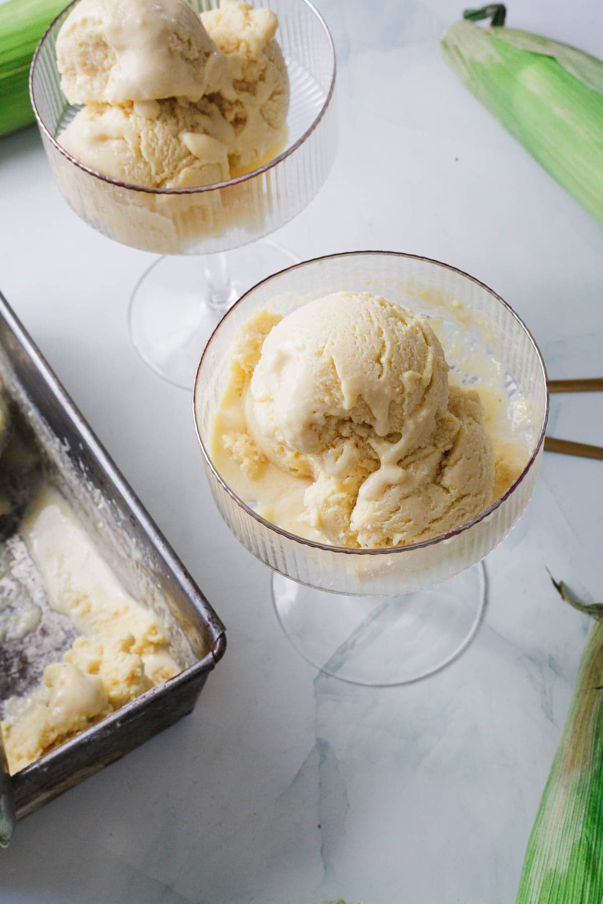 Two glass bowls filled with scoops of yellow Corn Ice Cream sit on a white surface. Nearby, a metal container with more creamy Corn Ice Cream and two ears of corn, one partially husked, complete the scene.