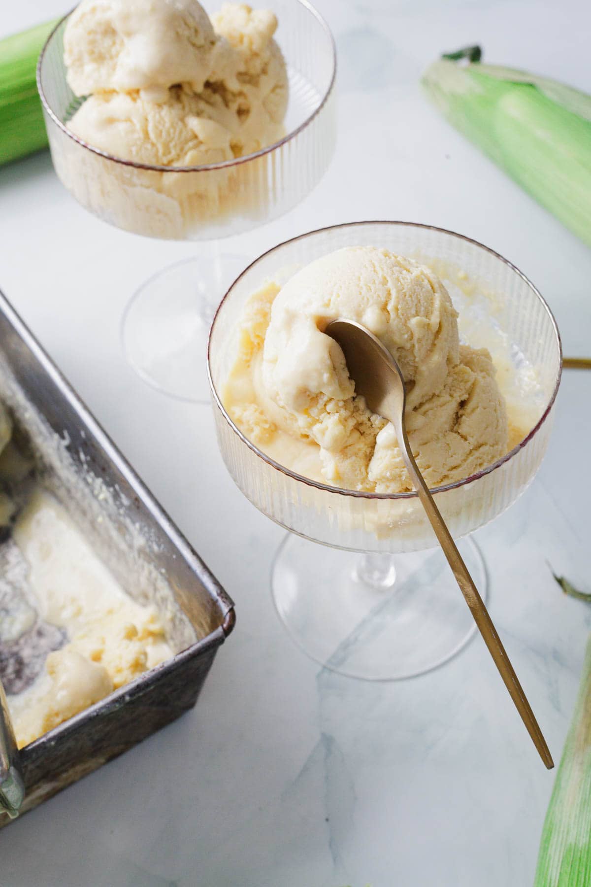 Two glass dessert cups each containing scoops of corn ice cream, with a spoon resting in one of them. A metal container with leftover ice cream and pieces of corn on the cob are also visible on a white surface.