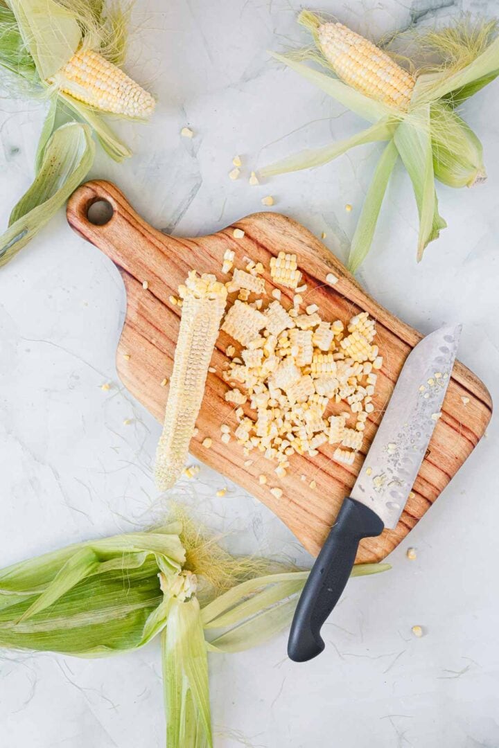 A wooden cutting board holds a partly stripped ear of corn, corn kernels, and a knife with a black handle—perfect prep for making Corn Ice Cream. Four more husked ears of corn rest on a light-colored surface around the board.