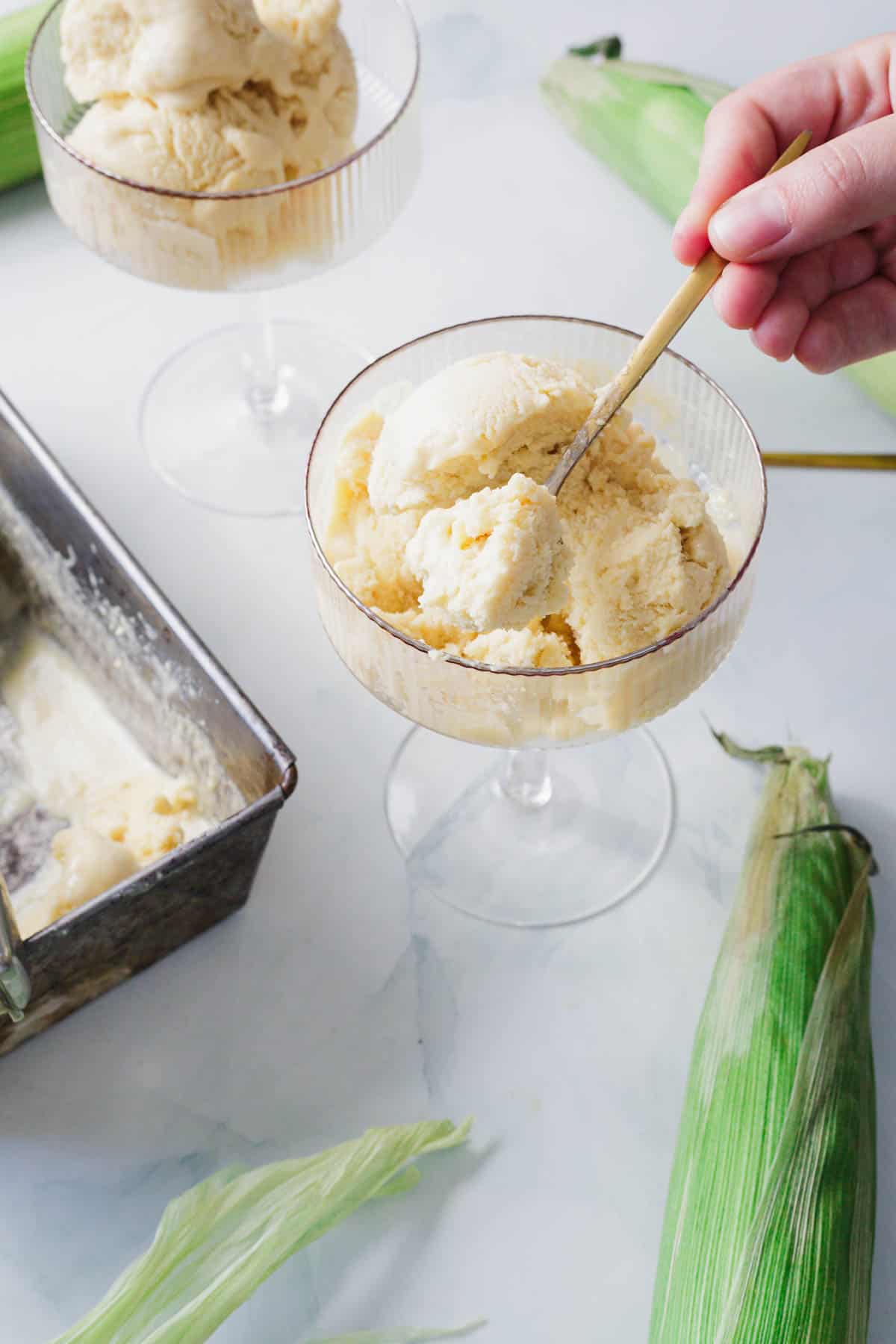 A hand holding a spoon scoops pale yellow Corn Ice Cream from a glass dish. Nearby, another dish with ice cream, a metal container filled with more Corn Ice Cream, and ears of corn with husks rest on a white surface.