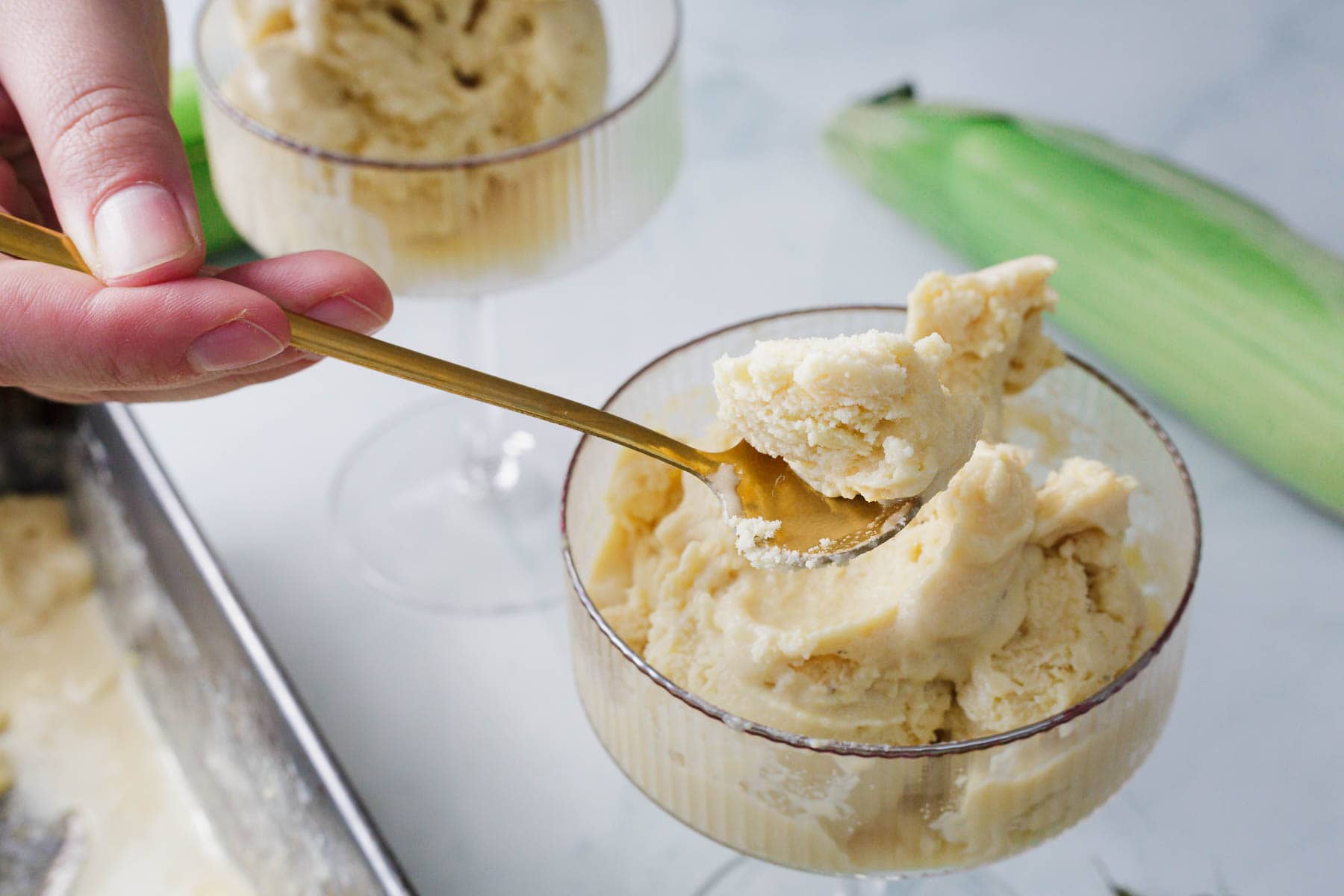 A hand holds a gold spoon, scooping light yellow Corn Ice Cream from a glass dessert dish. Another filled dish and an ear of corn are visible in the background on a white surface.