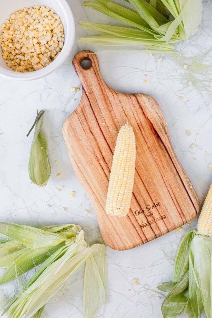 An ear of corn sits on a wooden cutting board surrounded by corn husks. A white bowl filled with kernels—perfect for making Corn Ice Cream—rests nearby on a marbled surface, with corn silk and husks scattered around.