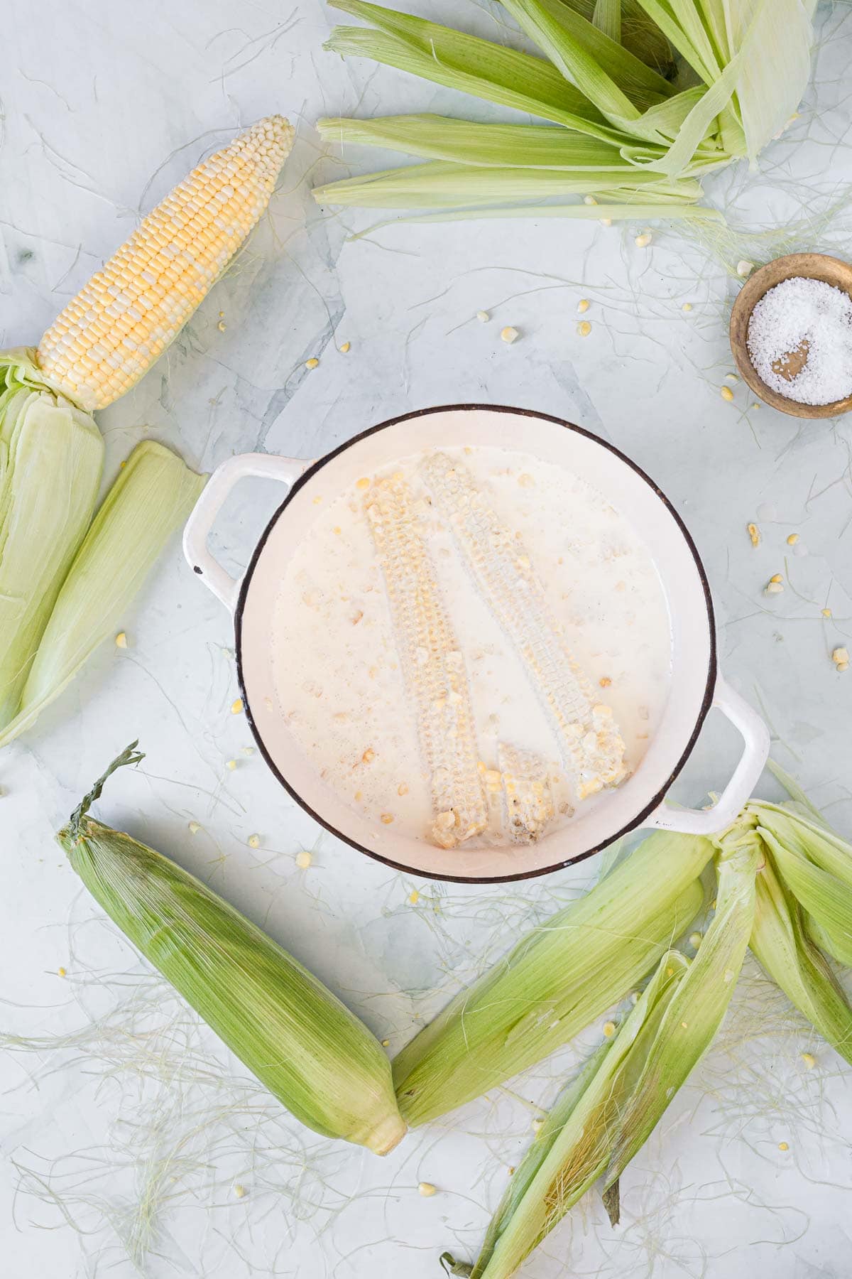 A white pot with soaked corn cobs and kernels in liquid, perfect for making Corn Ice Cream, sits on a light surface surrounded by fresh corn on the cob, corn husks, and a small bowl of salt.