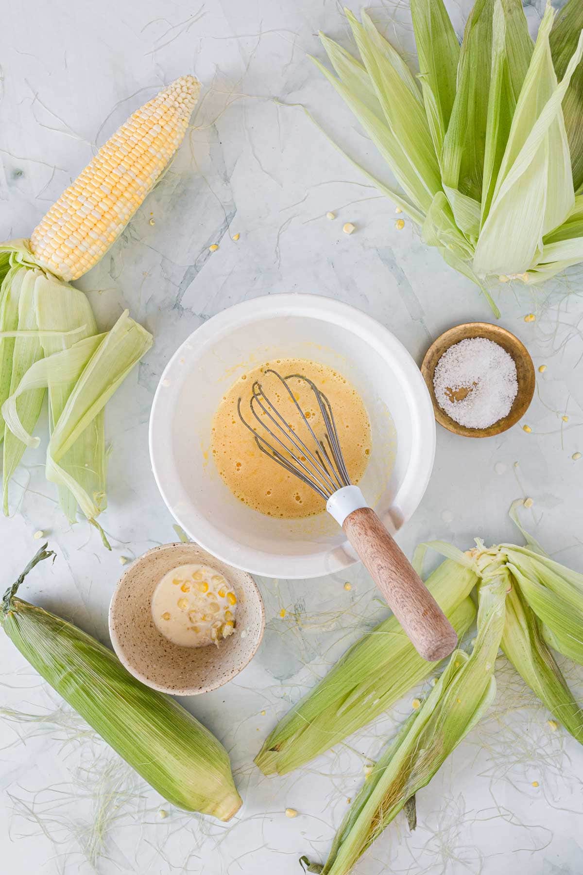A bowl with a whisk sits on a marble surface surrounded by corn on the cob with husks, a bowl of butter, and a small bowl of salt—classic ingredients for making Corn Ice Cream. Corn kernels and husks are scattered around.