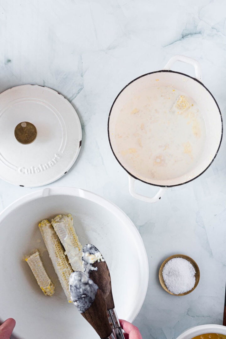 A person uses tongs to hold corn cobs over a white bowl. Nearby, a Dutch oven with creamy liquid hints at homemade Corn Ice Cream, with cobs inside, a matching lid, and a small bowl of salt on a light-colored surface.