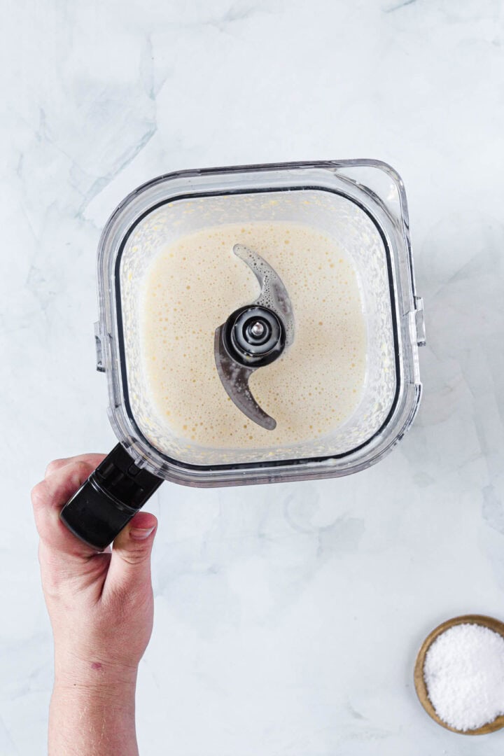 A hand holding the handle of a blender filled with a creamy, light beige corn ice cream mixture. A small bowl containing white powder sits on the surface nearby.