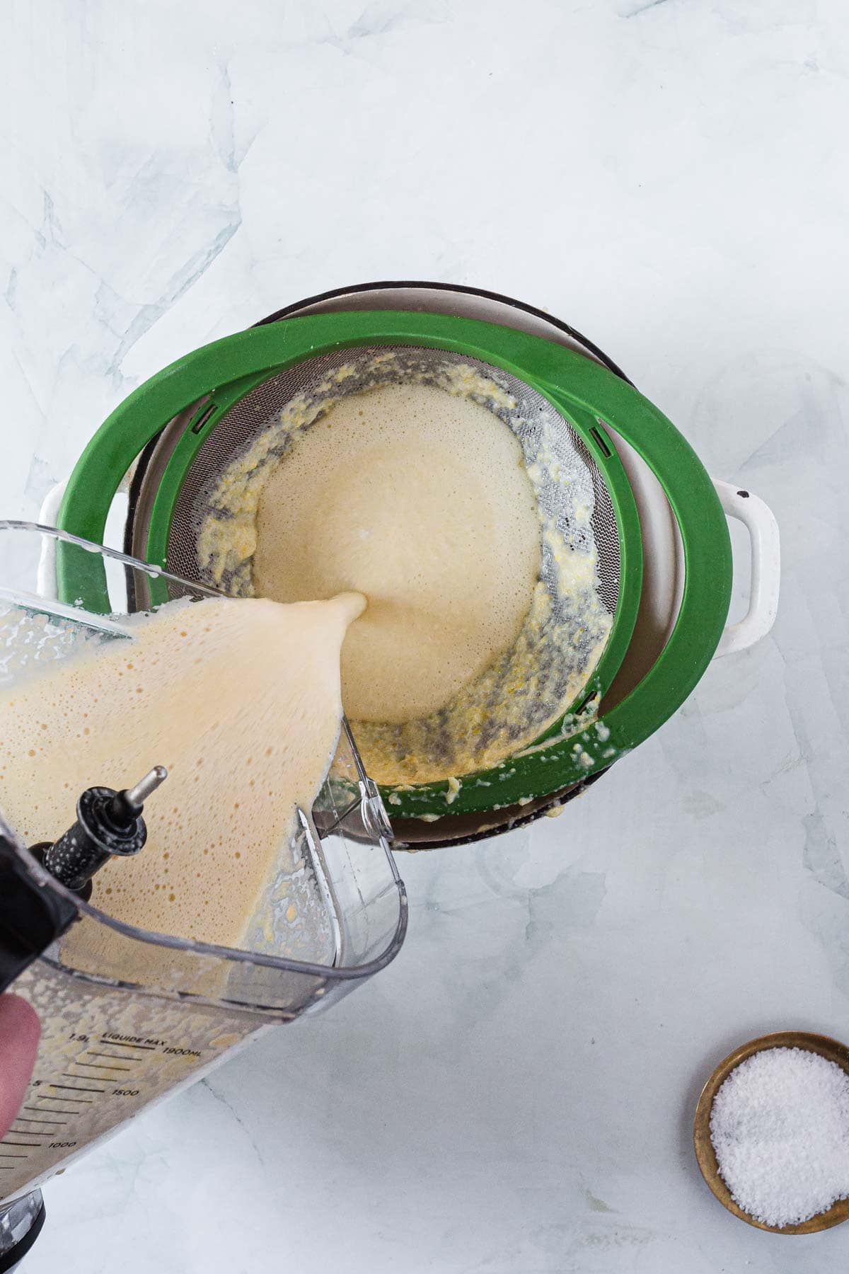 A person pours a blended, frothy corn ice cream mixture from a blender into a green mesh strainer set over a white bowl. A small bowl of salt sits on the right side of the image on a light surface.