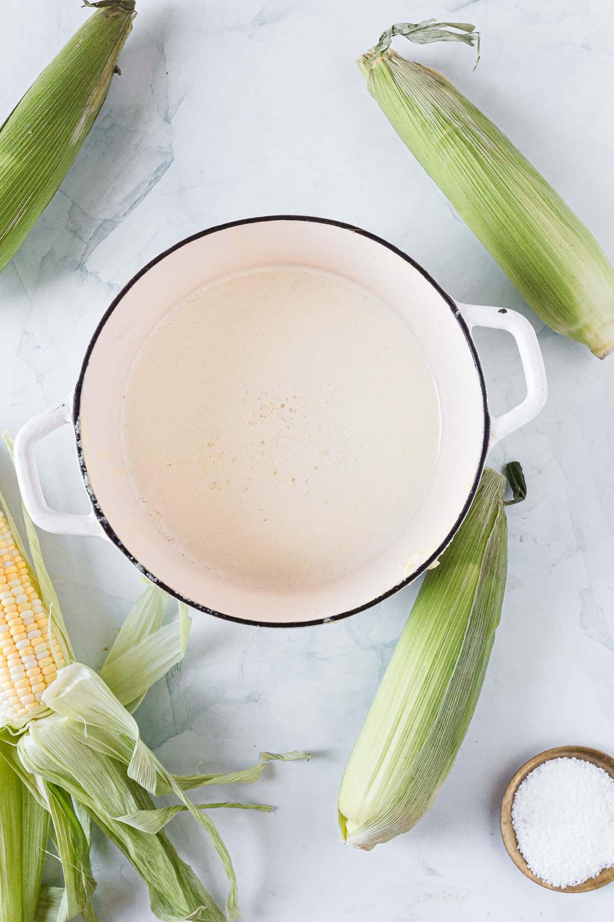 A white pot with creamy Corn Ice Cream inside is placed on a light surface, surrounded by fresh corn on the cob, some with husks partially removed, and a small bowl of salt.