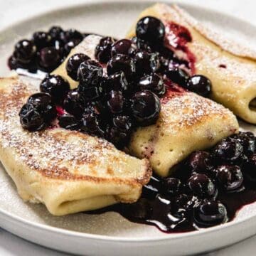 Three rolled crepes are served on a white plate, topped with powdered sugar and a generous portion of cooked blueberries in syrup. The dish sits on a light-colored surface.