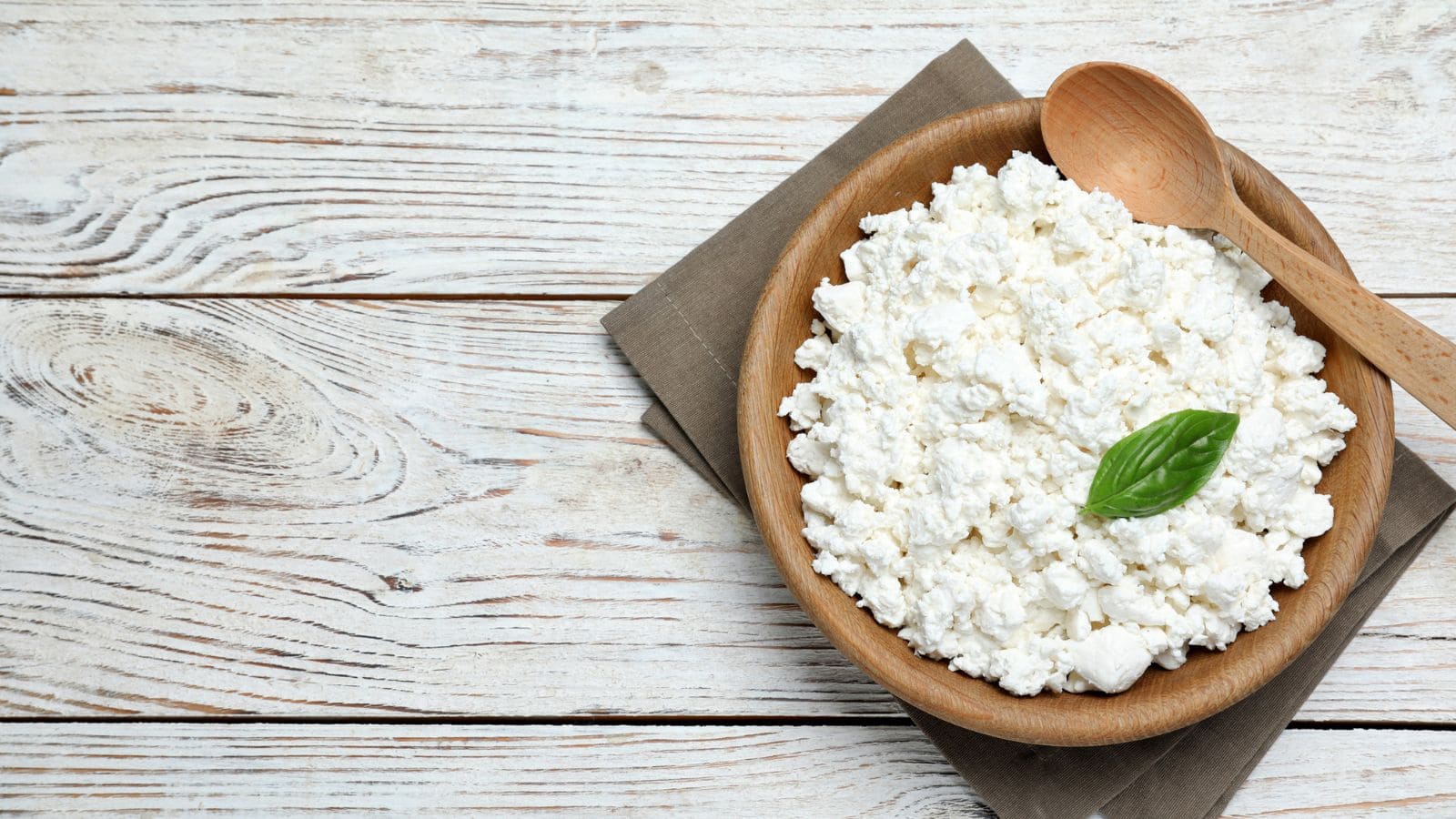 A wooden bowl filled with cottage cheese sits on a folded brown napkin, topped with a basil leaf. A wooden spoon rests on the bowl. The background is a whitewashed wooden surface.