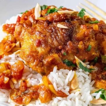 A plate of white rice topped with a serving of tomato-based stew with pieces of chicken, bell peppers, and garnished with sliced almonds and chopped herbs. A gold-colored fork rests on the plate.