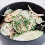 A black bowl filled with Creamy Hungarian Cucumber Salad, featuring thin cucumber slices, red onion, fresh dill, ground black pepper, and a light dressing. A fork is partially visible at the side of the bowl.