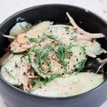 A black bowl filled with Creamy Hungarian Cucumber Salad, featuring thin cucumber slices, red onion, fresh dill, ground black pepper, and a light dressing. A fork is partially visible at the side of the bowl.