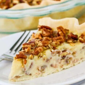 A slice of pie with a creamy filling and chopped pecans on top sits on a white plate with a fork. The rest of the pie is visible in a glass pie dish in the background.